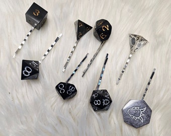 May include: A set of nine black and silver dice hair clips. The dice are in various shapes and sizes, including a d4, d6, d8, d10, d12, and d20. The hair clips are silver and have a simple design.