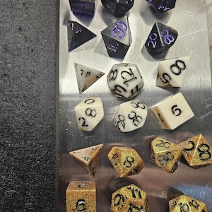 May include: A collection of polyhedral dice in various shapes and colors. Some dice are black with purple numbering, while others are white with black numbers. Several dice have a gold glitter finish with black numbering. The dice are arranged on a silver surface.