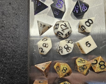 May include: A selection of multi-sided dice in different shapes and colours. Some are black with purple numerals, others are white with black numbers. Several dice have a gold glitter finish with black numbering. The dice are arranged on a silver surface.