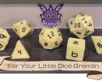 May include: A set of yellow crocheted dice with black numbers. The dice are arranged on a wooden surface with a purple crocheted dragon head in the background. The text "For Your Little Dice Gremlin" is written on a brown banner.