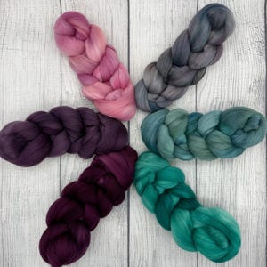 May include: Five braids of colorful wool roving in shades of pink, gray, teal, and purple arranged in a starburst pattern. The wool is soft and fluffy, perfect for fiber art projects like felting and spinning. The background is a light wood grain.