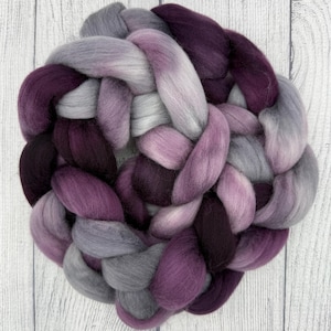 May include: A braided roving of wool in shades of purple, lavender, and gray. The fiber is arranged in a circular pattern, with the colors blending together. The wool is soft and fluffy, ideal for various fiber art projects.
