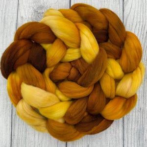 May include: A close-up of a braided bundle of wool roving in shades of yellow and brown. The wool is arranged in a circular pattern, with the colors blending together. The texture of the wool is soft and fluffy, ideal for fiber art projects.