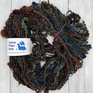 May include: A skein of handspun yarn in a mix of dark colors, including brown, blue, and green, with strands of silver thread. The label reads "Ajem Fiber Arts Handspun Yarn" with a camel logo.