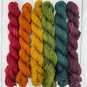 May include: Six skeins of twisted yarn arranged vertically in a gradient of colors including deep red, bright orange, golden yellow, olive green, dark teal, and rich plum purple, each featuring a multi-ply twisted texture and consistent thickness.