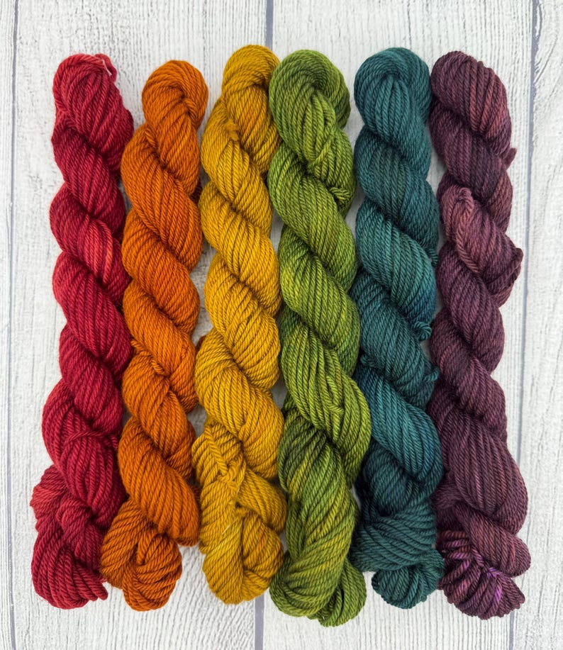 May include: Six skeins of twisted yarn arranged vertically in a gradient of colors including deep red, bright orange, golden yellow, olive green, dark teal, and rich plum purple, each featuring a multi-ply twisted texture and consistent thickness.