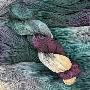 May include: A twisted skein of yarn featuring a gradient color transition. The colors shift from deep forest green and teal to dark purple, transitioning into a soft beige at one end. The yarn has a smooth, plied texture throughout the skein.