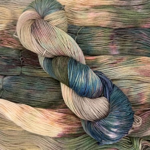 May include: A skein of hand-dyed yarn featuring a variegated color palette of muted earth tones, including shades of beige, olive green, deep forest green, and soft hints of dusty rose, twisted into a classic figure-eight shape for display.