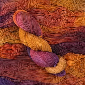 May include: A skein of hand-dyed yarn featuring a variegated color palette of deep magenta, burnt orange, and golden yellow. The twisted yarn rests atop a background of loose, unspun strands of the same multi-colored fiber, creating a textured surface.