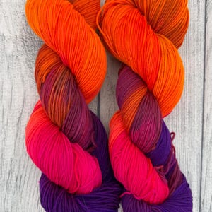 May include: Two skeins of hand-dyed yarn featuring a vibrant gradient color transition. The color palette shifts from a bright orange at the top, blending into deep magenta, and finishing with a rich purple at the bottom of each twisted hank.