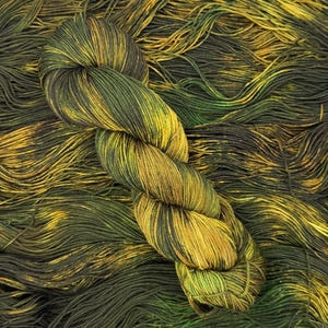 May include: A skein of yarn featuring a variegated color palette of deep forest green, olive, and bright golden yellow. The yarn is twisted into a compact bundle and rests atop a loose, spread-out layer of the same multi-colored fiber strands.