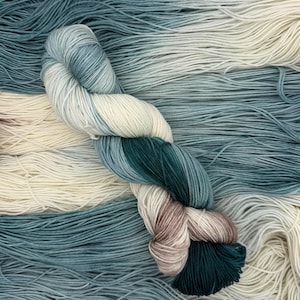 May include: A twisted skein of yarn featuring a gradient color palette that transitions from deep teal and muted blue to creamy off-white and soft taupe. The yarn is displayed against a background of loose, unraveled strands of the same fiber.
