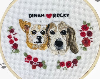 May include: Embroidered hoop art featuring two dogs with the names "DINAH" and "ROCKY" above them, separated by a red heart. The dogs are surrounded by a floral wreath of red and green embroidered details, set against a white fabric.