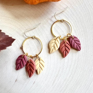 May include: Gold hoop earrings, each featuring three leaf charms in burgundy, rust, and cream. The earrings are presented on a light wood surface, creating a seasonal autumn aesthetic.