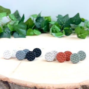 May include: A collection of round stud earrings in various colors, including white, gray, black, terracotta, and sage green. Each earring features a textured leaf pattern. The earrings are displayed on a wooden slice with green foliage in the background.