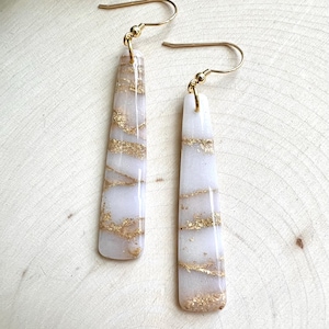 May include: A pair of rectangular dangle earrings. The earrings are white with gold-colored accents. The gold accents are in horizontal lines across the length of the earrings. The earrings have gold-colored hooks.