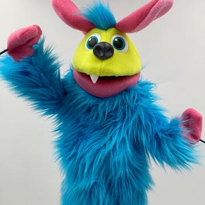 May include: A colorful puppet with a yellow face, blue fur, and pink accents. The puppet has large, pink ears, a black nose, and a wide mouth with a single white tooth. The puppet's arms are outstretched, and it appears to be made of soft, plush materials.