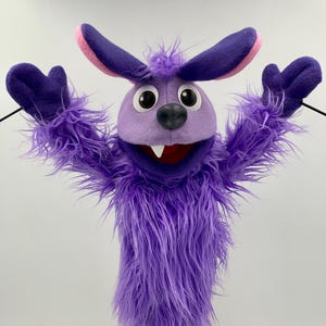 May include: A purple puppet with a furry body, large ears with pink inner ears, and a round head. The puppet has big, round eyes, a black nose, and a red mouth with two white teeth. The puppet's arms are raised, and it is mounted on a wooden stand.