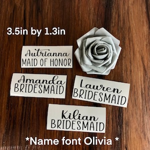 Peut inclure: &Eacute;tiquettes nominatives en vinyle blanc avec texte noir, dont "Autrianna MAID OF HONOR", "Amanda BRIDESMAID", "Lauren BRIDESMAID" et "Kilian BRIDESMAID". Une rose en tissu argent&eacute; est &eacute;galement pr&eacute;sente. Les &eacute;tiquettes mesurent 8,9 cm sur 3,3 cm.