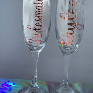 Peut inclure: Deux fl&ucirc;tes &agrave; champagne transparentes avec des lettres en or rose. L'une porte l'inscription "Bridesmaid" et l'autre "Kaylee". Les fl&ucirc;tes sont remplies de papier de soie blanc et reposent sur une surface r&eacute;fl&eacute;chissante.