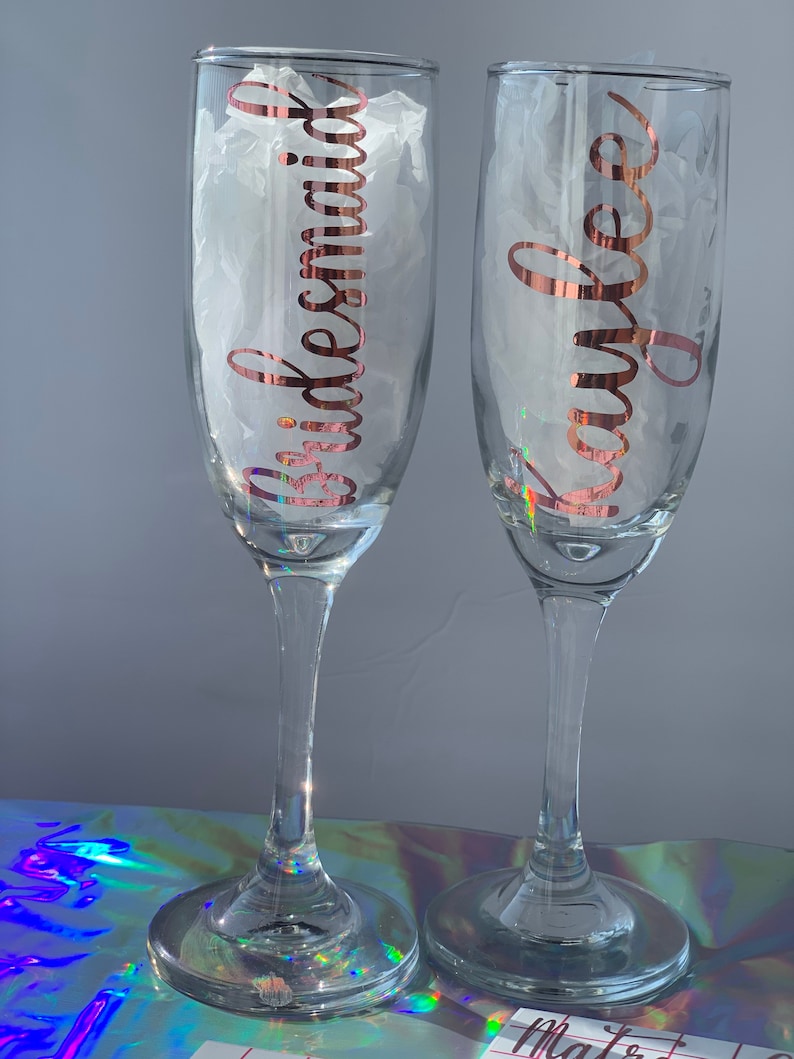Peut inclure: Deux fl&ucirc;tes &agrave; champagne transparentes avec des lettres en or rose. L'une porte l'inscription "Bridesmaid" et l'autre "Kaylee". Les fl&ucirc;tes sont remplies de papier de soie blanc et reposent sur une surface r&eacute;fl&eacute;chissante.