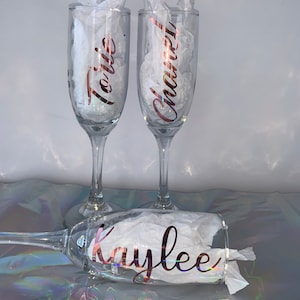 Peut inclure: Trois fl&ucirc;tes &agrave; champagne transparentes avec des noms en script or rose : "Torie", "Chanel" et "Kaylee". Chaque verre est rempli de papier de soie blanc. Les verres sont sur une surface iris&eacute;e et r&eacute;fl&eacute;chissante.