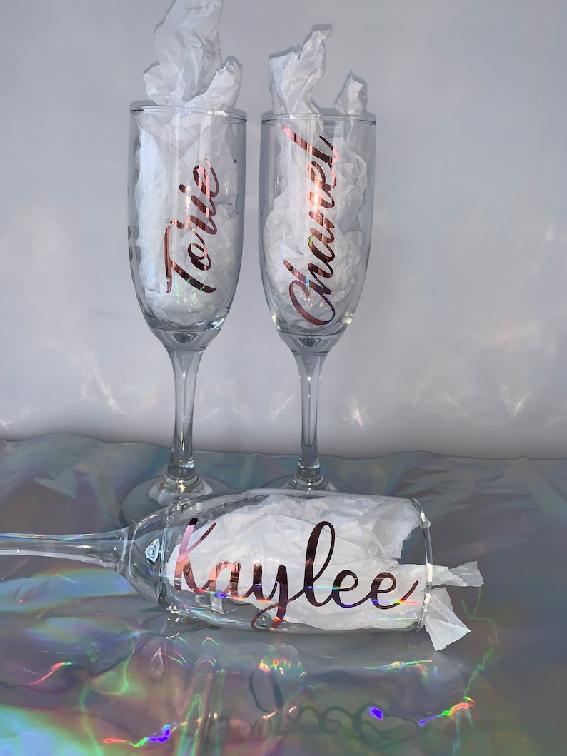 Peut inclure: Trois fl&ucirc;tes &agrave; champagne transparentes avec des noms en script or rose : "Torie", "Chanel" et "Kaylee". Chaque verre est rempli de papier de soie blanc. Les verres sont sur une surface iris&eacute;e et r&eacute;fl&eacute;chissante.