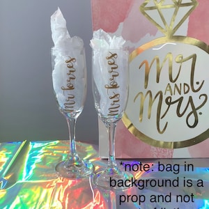 Peut inclure: Deux fl&ucirc;tes &agrave; champagne transparentes avec l'inscription dor&eacute;e "Mr Torres" et "Mrs Torres". Chaque fl&ucirc;te est remplie de papier de soie blanc. Un sac cadeau avec le texte "Mr and Mrs" est &agrave; l'arri&egrave;re-plan.