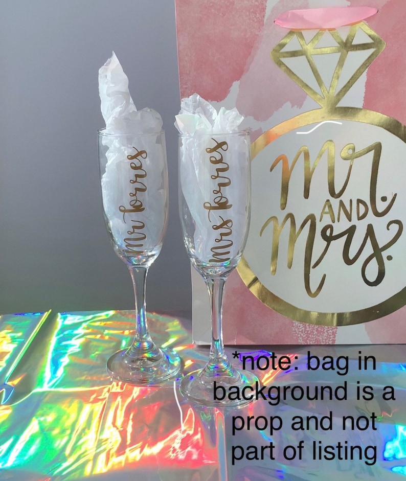 Peut inclure: Deux fl&ucirc;tes &agrave; champagne transparentes avec l'inscription dor&eacute;e "Mr Torres" et "Mrs Torres". Chaque fl&ucirc;te est remplie de papier de soie blanc. Un sac cadeau avec le texte "Mr and Mrs" est &agrave; l'arri&egrave;re-plan.