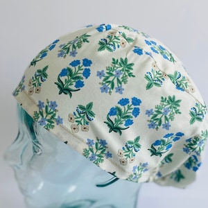 May include: A white scrub cap with a blue and green floral pattern. The cap is on a glass mannequin head.
