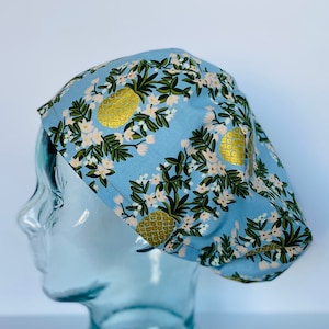 May include: A light blue scrub hat with a pineapple and floral print. The hat is on a glass mannequin head.
