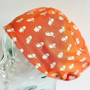 May include: Orange Halloween-themed scrub cap with a repeating pattern of white cartoon ghosts. The cap is made of fabric and is designed to cover the head. The background is white.
