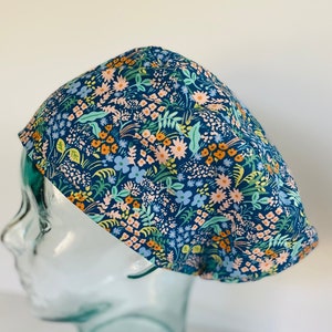 May include: A navy blue surgical scrub cap with a floral pattern. The cap features a design of small flowers and leaves in shades of peach, light blue, and green. The cap is displayed on a clear glass head form.