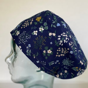 May include: A navy blue surgical scrub cap with a floral pattern. The cap features a repeating pattern of white, yellow, and blue flowers with green leaves. The cap is made of a soft, lightweight fabric.