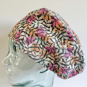 May include: A colorful surgical scrub cap featuring a repeating pattern of leaves in pink, orange, and white on a black background. The cap is made of fabric and is designed to cover the head.