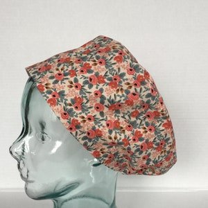 May include: A floral patterned scrub cap featuring a cream background with small pink roses and gray leaves. The cap is displayed on a clear glass head form, showcasing its shape and design. The fabric appears to be cotton.