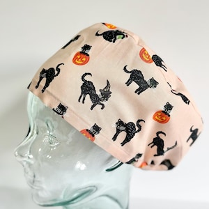 May include: A light pink scrub cap featuring a repeating pattern of black cats, some wearing witch hats, and orange jack-o'-lantern pumpkins. The fabric has a soft, matte finish and is shaped to fit snugly around the head with a rounded crown.