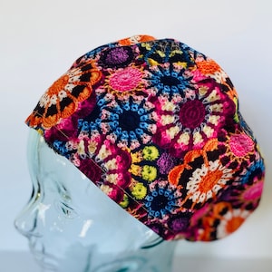 May include: A colorful surgical scrub hat with a crocheted pattern in shades of pink, orange, blue, green, and white on a dark background.