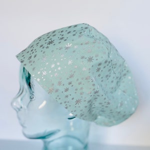 May include: A light blue surgical scrub cap with a silver snowflake pattern. The cap is made of a soft fabric and is designed to cover the hair during medical procedures. The cap is shown on a glass head.
