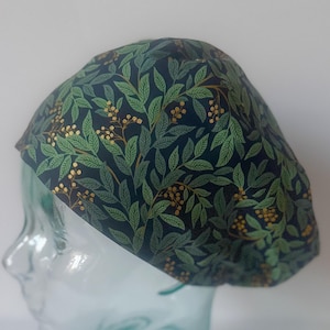 May include: A dark blue surgical scrub cap with a green and gold leaf and berry pattern. The cap is made of fabric and is designed to cover the head. The cap is shown on a glass mannequin head.