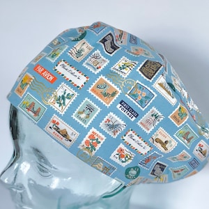 May include: A blue scrub hat with a pattern of colorful postage stamps from around the world. The stamps feature images of animals, flowers, and landmarks. The text "Blue Dolphin" and "Par Avion" are visible on the stamps.