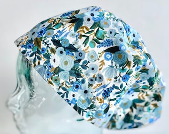 Puede incluir: Un gorro quirúrgico con estampado floral azul. El gorro presenta un fondo blanco con diseños florales azules, turquesas y dorados. El gorro se muestra sobre una cabeza de cristal transparente sobre un fondo blanco.