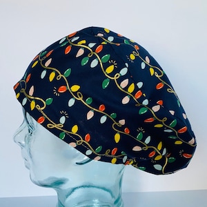 May include: A navy blue surgical scrub cap with a repeating pattern of colorful Christmas lights and gold-colored string. The lights are in shades of red, yellow, green, and pink. The cap is designed to fit comfortably and securely.