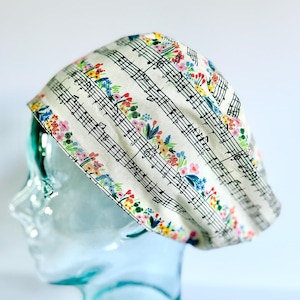 May include: A white scrub hat with a floral and music note pattern. The hat is on a glass mannequin head.