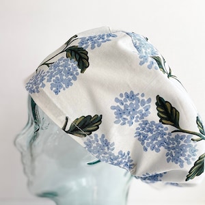 May include: A white fabric hat with a blue floral pattern featuring clusters of blue flowers and green leaves. The hat is designed to fit the head and is likely for medical or culinary use.