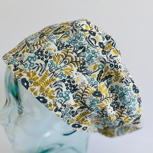 May include: A white scrub cap with a floral pattern in shades of blue, green, and gold. The cap is made of cotton and has a tie closure.