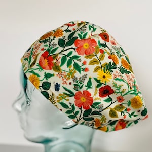 May include: A white scrub hat with a floral print featuring red, yellow, and green flowers. The hat is on a glass mannequin head.