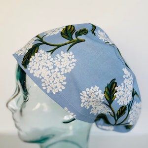 May include: A light blue surgical scrub cap with a floral pattern. The cap features white hydrangea flowers with green leaves and stems. The cap is made of fabric and is designed to cover the head.