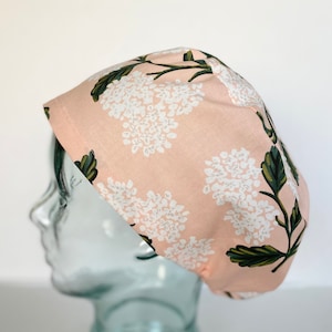 May include: A pink surgical scrub cap with a white floral pattern and green leaves. The cap is made of fabric and is designed to cover the hair during medical procedures. The cap is shown on a glass head.