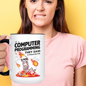 Op de afbeelding: Een witte koffiemok met een zwarte rand en handvat. De mok heeft een cartoon-skelet in vlammen met de tekst "Computer Programming They Said It Would Be Fun."